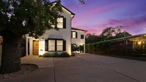 A two-story house with a well-lit front entrance, a large tree on the left, and a driveway leading to the house. - 🏝️LUX Hot Tub +Chiller Near DT: Spa Baths, Yard Games, Arcade, Parking, SLEEPS 18 (Austin)