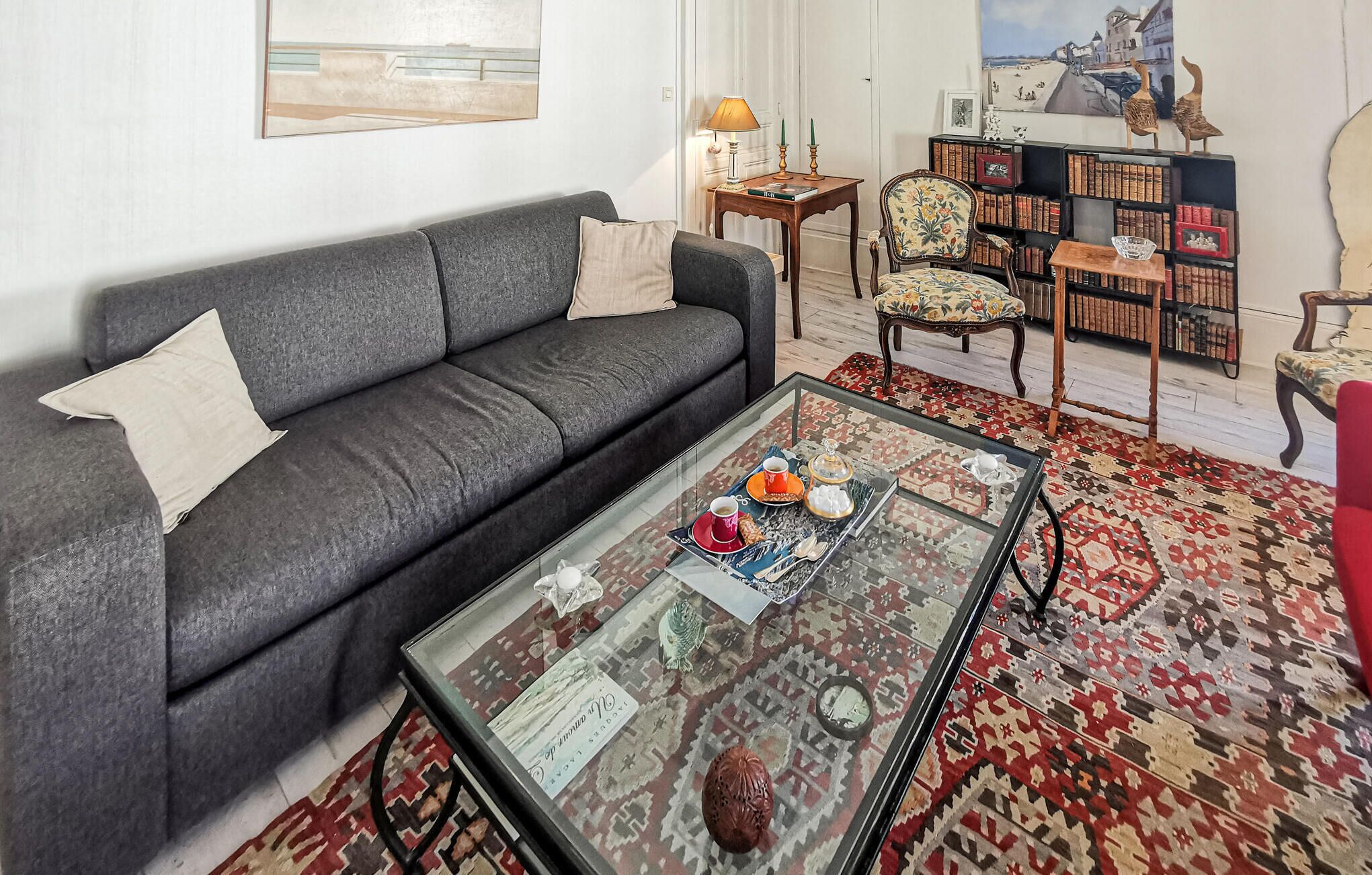 A living room with a grey sofa, a glass coffee table with decorative items, a patterned rug, and a bookshelf with books and decorative items.