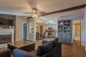 A spacious living room with a fireplace, a ceiling fan, and a dining area with a kitchen in the background.