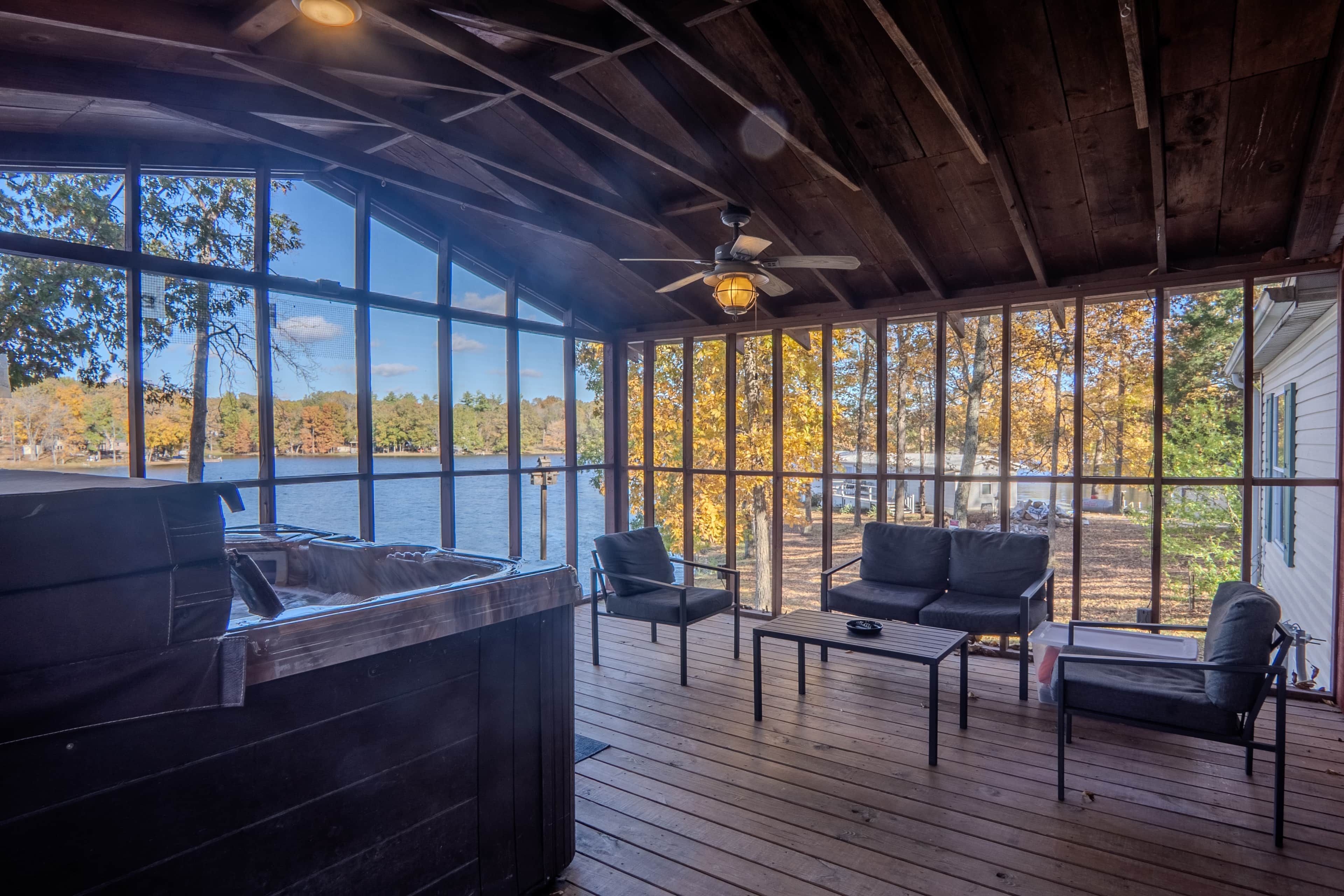 Une terrasse extérieure couverte, dotée d’un jacuzzi, d’un espace salon et de grandes fenêtres offrant une vue sur un lac et les couleurs automnales des arbres.