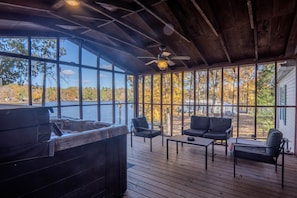 Terraza exterior cubierta con jacuzzi, zona de estar y amplios ventanales con vistas a un lago y follaje otoñal.