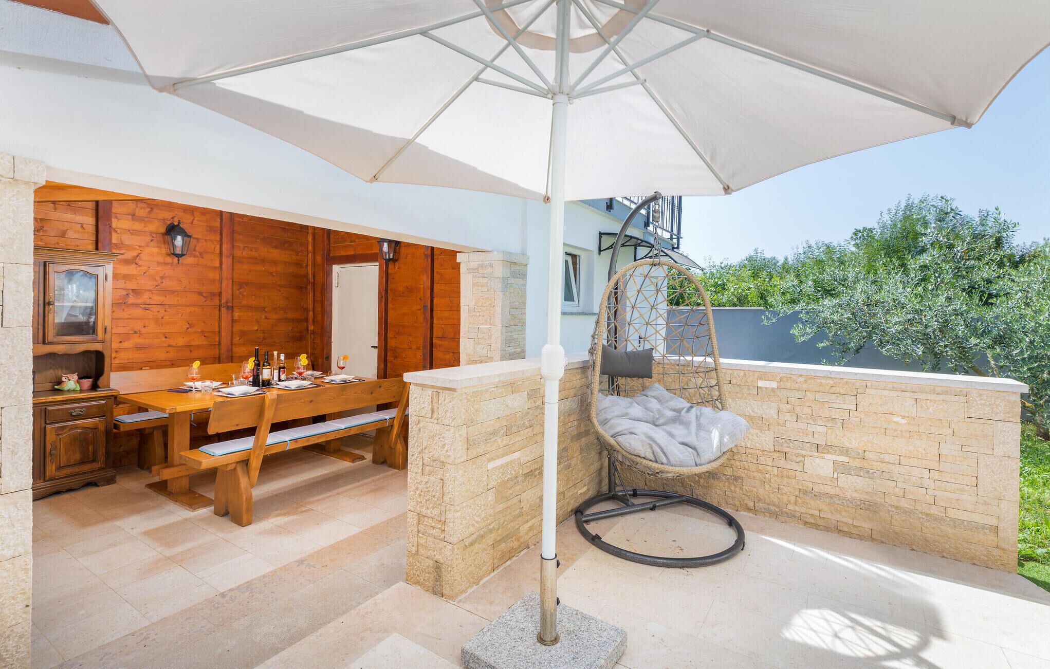 A patio with a wooden table set for dining, a hanging chair, and a large umbrella providing shade.