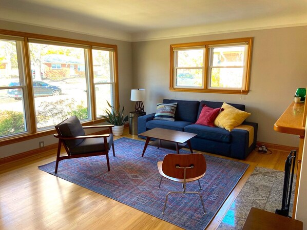 A living room with a blue sofa, two armchairs, a coffee table, and a rug. There are two windows with a view of the outside. - Cozy 4-Bedroom Family Home - Short Walk to Michigan Stadium! (Ann Arbor)