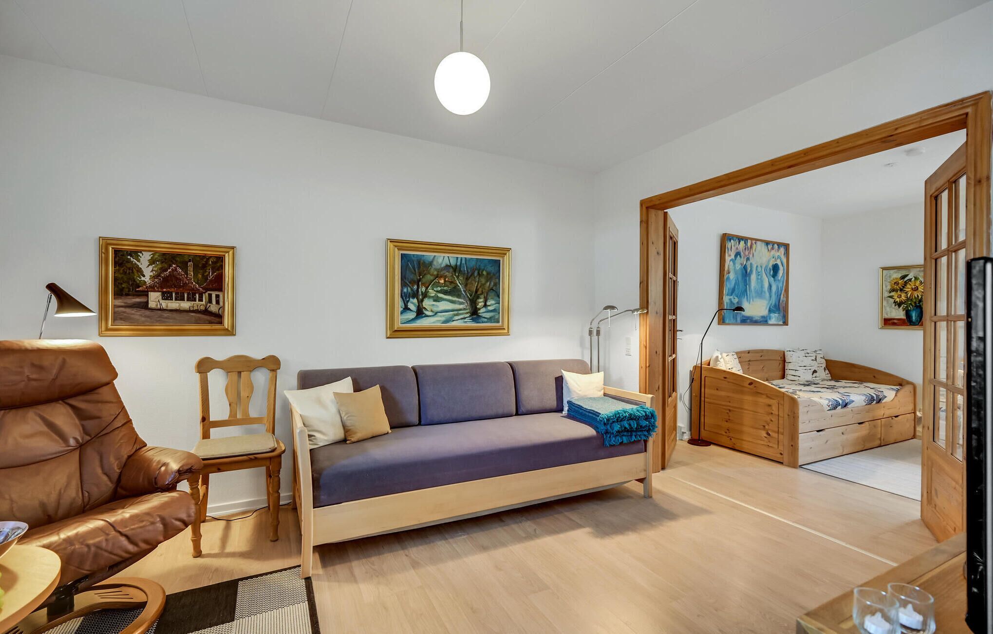 Een moderne woonkamer met een bank, fauteuil en een houten bed. Er hangen schilderijen aan de muren en er staat een lamp op de vloer.