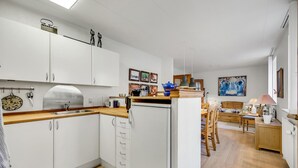 Ett modernt kök med vita skåp, en fläktkåpa i rostfritt stål och en bänkskiva i trä. Det finns en matplats med ett träbord och stolar, och i bakgrunden syns en säng med en sänggavel i trä.