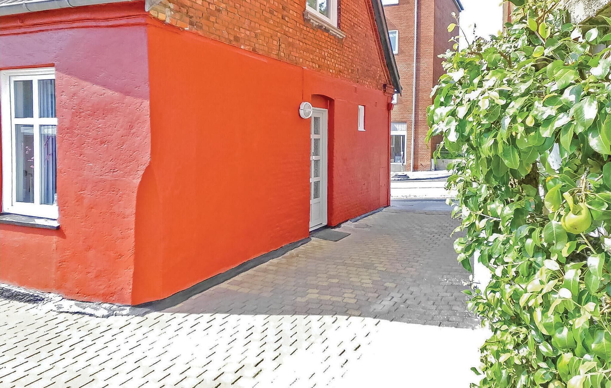 A red brick building with a white door and windows, a paved walkway, and a green hedge.