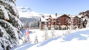 Une station de montagne enneigée, avec des bâtiments à plusieurs étages, des pistes de ski et un ciel bleu dégagé.
