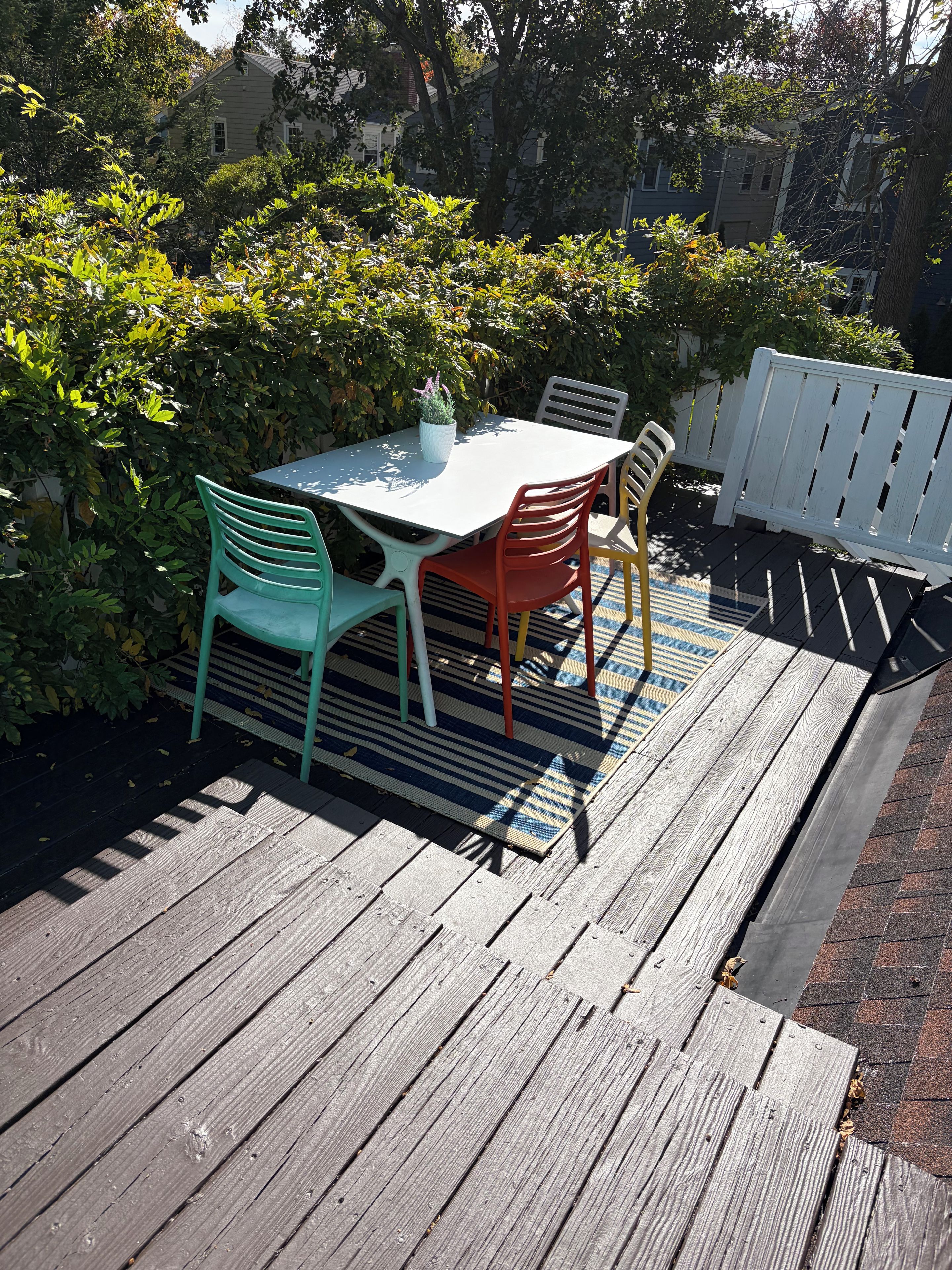 Una terraza de madera con una mesa y sillas, rodeada de vegetación y una valla blanca.