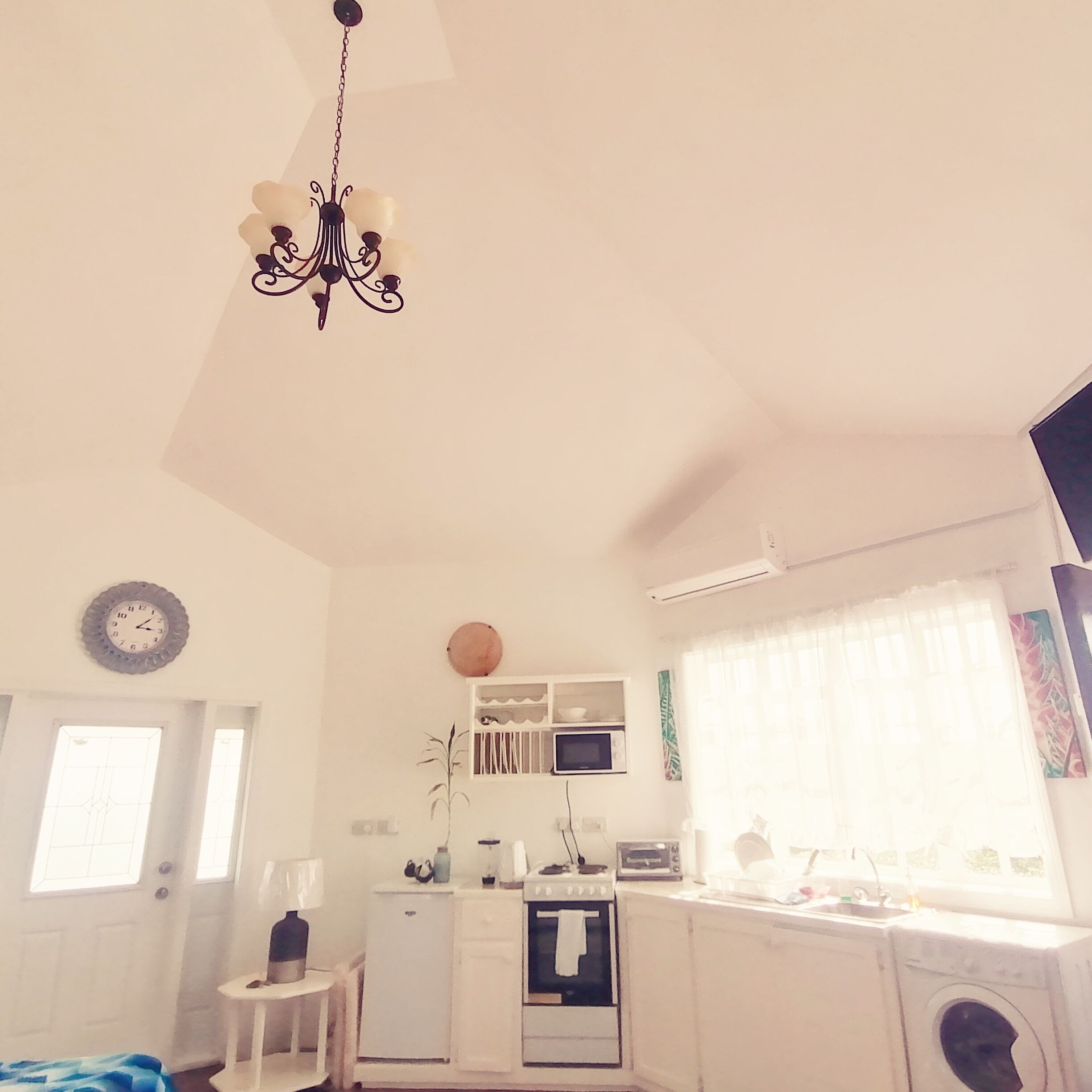 A compact kitchen with a microwave, oven, and sink. There is a small table with a lamp, a clock on the wall, and a ceiling light fixture.