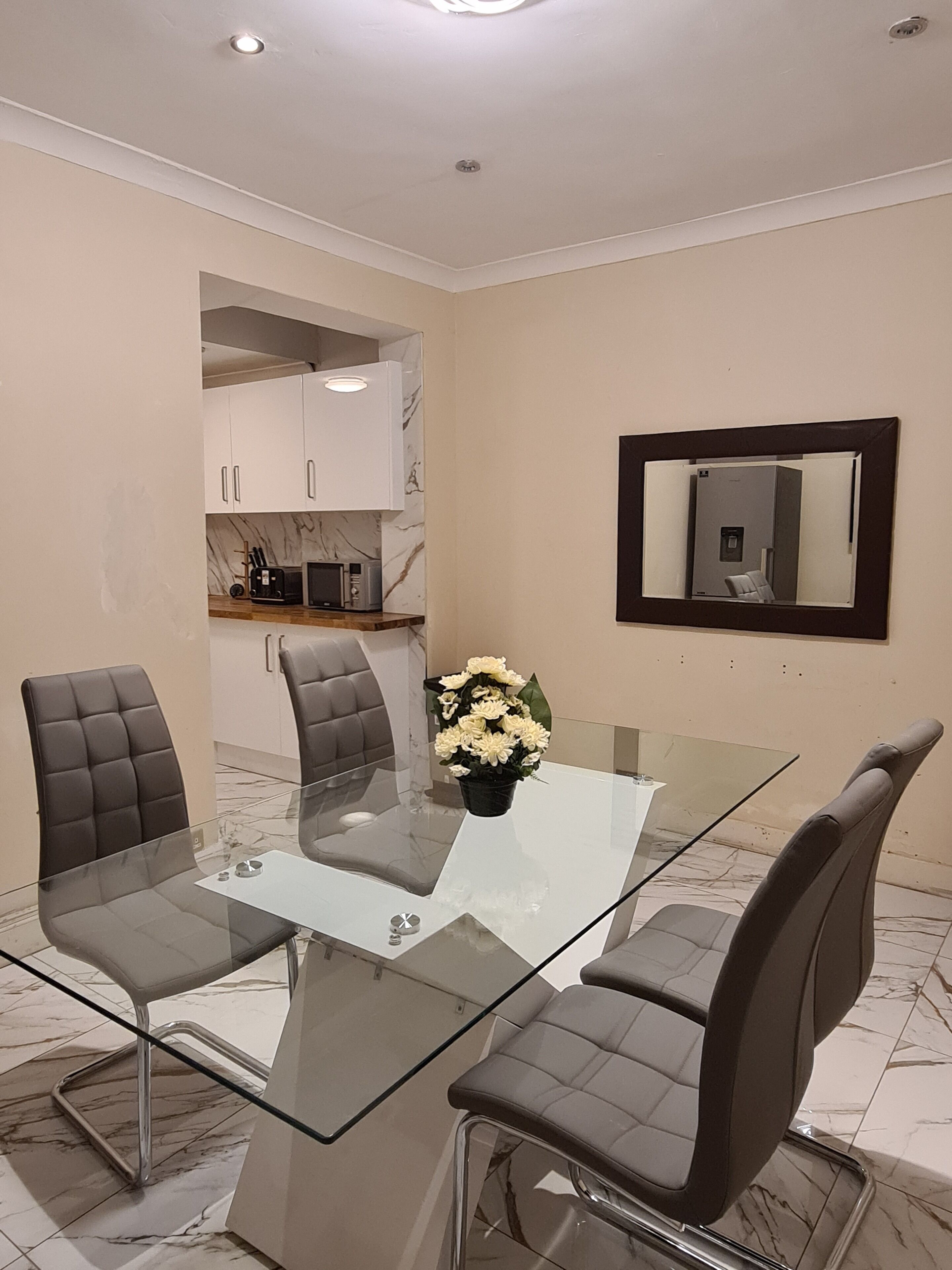 A modern dining area with a glass table and four chairs, a vase of flowers, and a kitchen in the background.
