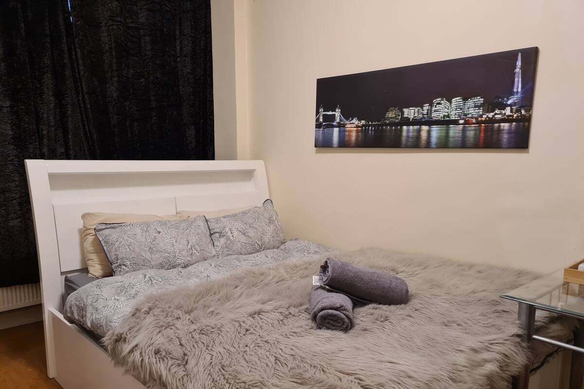 A neatly made bed with a fluffy grey blanket and pillows, a rolled towel, and a cityscape painting on the wall.