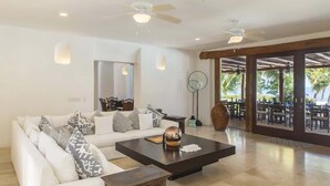 A spacious living area with a white sofa, a dark coffee table, and a wooden side table. There is a large mirror on the wall and a ceiling fan. The room opens to a covered outdoor seating area with tables and chairs.