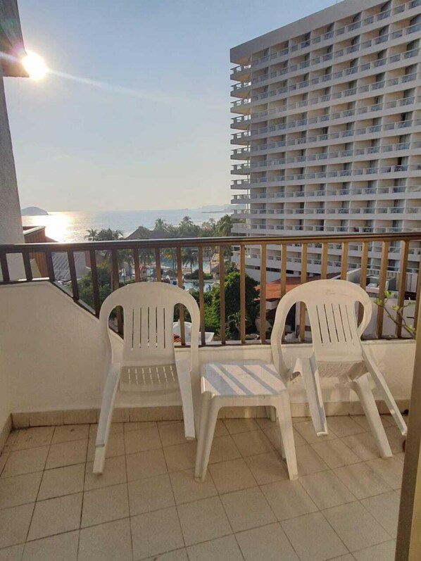 A balcony with two white plastic chairs, a tiled floor, and a view of a high-rise building and the ocean. - Condominio 01 recamara Enna Inn Ixtapa (Ixtapa)