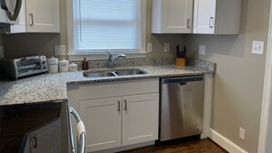 A compact kitchen with white cabinets, a granite countertop, a built-in dishwasher, and a window with blinds. - Delightful house just 10 minutes from uptown (Charlotte)