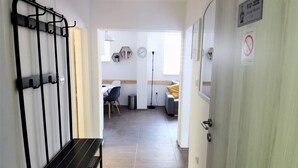 Un couloir avec un banc, une porte avec une pancarte et une vue sur une pièce où se trouvent un lit et une chaise.