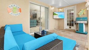 Ein Zimmer mit einem blauen Sofa, einer Glastür, die auf einen Balkon führt, einem an der Wand befestigten Fernseher und einem Schild im Strandhausstil.