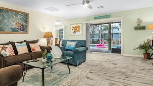 Ein Wohnzimmer mit braunem Sofa, einem türkisfarbenen Sessel und einem Glastisch. Eine Deckenventilator, ein Gemälde einer Meeresschildkröte und ein Schild mit der Aufschrift 'The Beach' sind zu sehen.