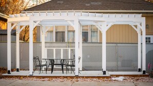 A covered patio with a table and chairs, surrounded by a white fence and a chain-link fence. - Grand View · Madison Place (Apt #1) Grand View (Ogden)