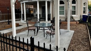A covered patio with a table and chairs, a white fence, and a brick building in the background. - Grand View · Madison Place (Apt #1) Grand View (Ogden)