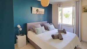 Une chambre à coucher avec un lit, des oreillers, une table de chevet, un vase de fleurs et une fenêtre avec des rideaux.