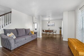 A modern living room with a grey sofa, a dining area with a table and chairs, and a staircase with white railings.