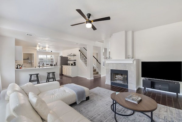 A spacious living room with a white sofa, a fireplace, a flat-screen TV, and a ceiling fan.