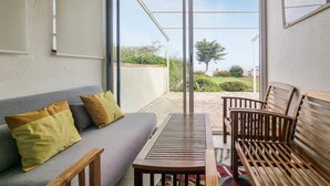 Une pièce comprenant un canapé, une table en bois et un banc. Une porte-fenêtre coulissante donne sur un espace extérieur offrant une vue sur la verdure et un bâtiment situé au loin.