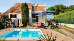 Une piscine entourée de chaises longues et de parasols, elle-même bordée d’un jardin aménagé et d’une maison dont le toit est recouvert de tuiles.