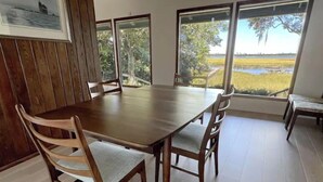 A dining area with a wooden table and chairs, large windows with a view of a natural landscape. - MCM Magic on the Marsh (Mount Pleasant)