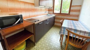 Kompakti keittiö, jossa on mikroaaltouuni, pesuallas ja puiset kaapit. Siellä on ikkuna, josta tulee luonnonvaloa, sekä pöytä ja tuolit.