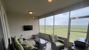A spacious living area with a large window offering a view of the countryside. There is a television mounted on the wall, a small table, and two wicker chairs with cushions. - Feet in the Sand Home- 13 Val (Laguna Vista)