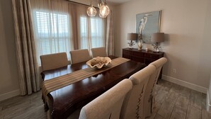 A dining room with a wooden table, beige chairs, a chandelier, and a painting of a bird on the wall. - Feet in the Sand Home- 13 Val (Laguna Vista)