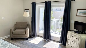 A room with a beige armchair, a floor lamp, a TV on a dresser, and a view of a white fence and building outside.