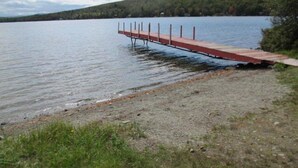 Un muelle de madera se extiende sobre un lago tranquilo, rodeado de orillas cubiertas de hierba y una colina boscosa al fondo.