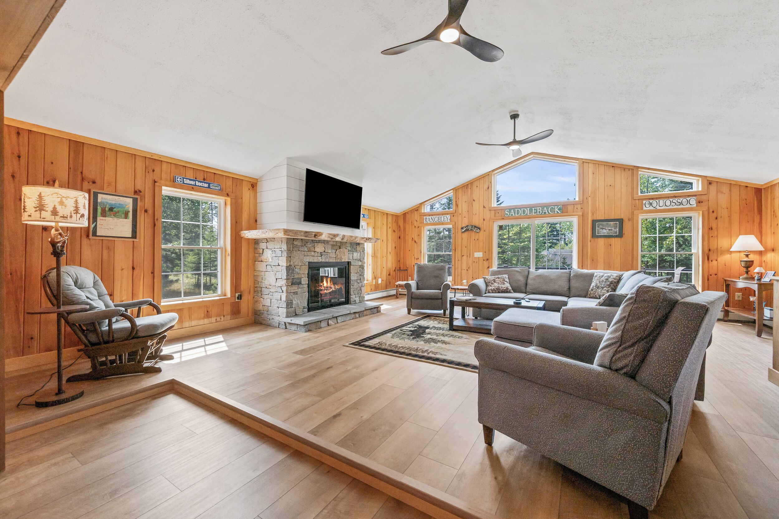 A spacious living room with a stone fireplace, wooden walls, and large windows.
