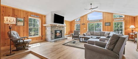 A spacious living room with a stone fireplace, wooden walls, and large windows.