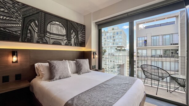 A modern hotel room with a large bed, a balcony with a view of the city, and a decorative wall piece above the bed.