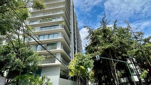 A multi-story building with balconies, surrounded by trees and a clear sky.