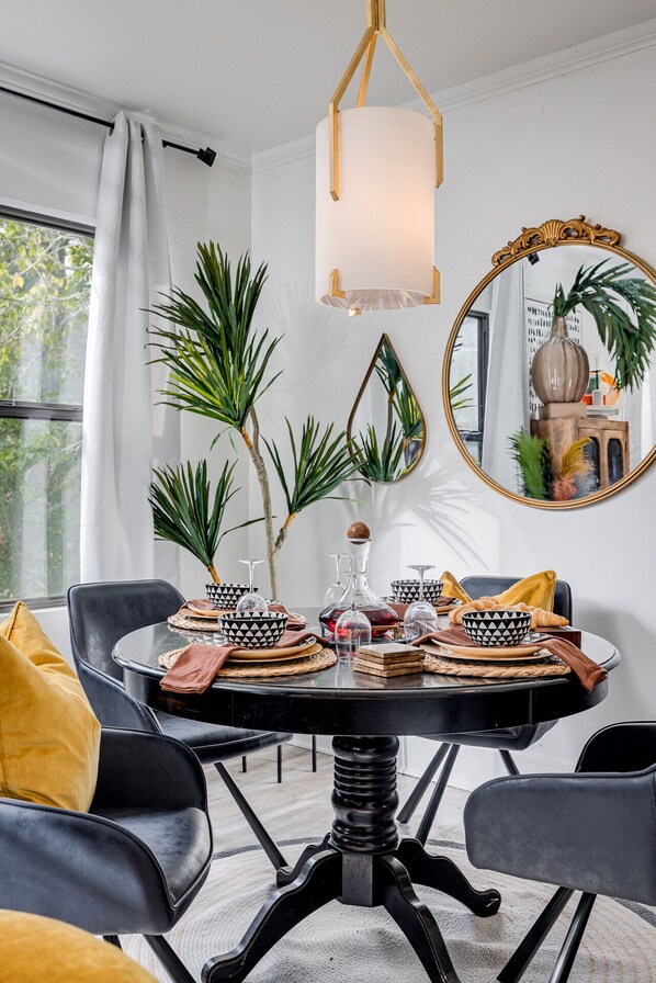 Een eetruimte met een ronde tafel, zwarte stoelen en een kroonluchter. Er staan planten, een spiegel en divers servies op tafel.