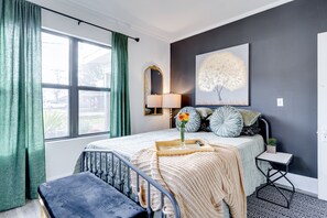 Een slaapkamer met een groot raam, een bed met een gebreide deken, een nachtkastje met een lamp en een spiegel aan de muur.