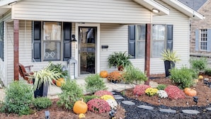 A single-story house with a front porch, a small garden with various plants and pumpkins, and a clear sky.