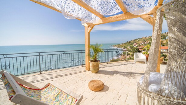A terrace with a view of the sea, a wicker chair, a potted plant, and a white chair.