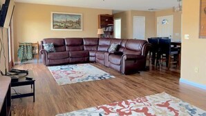 A living room with a sectional sofa, a ceiling fan, a painting on the wall, and a dining area with chairs and a table. - Walker's Refuge- large family home, close to river trails (Cody)