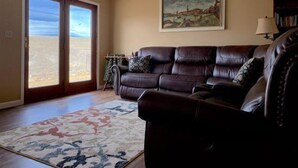 A living room with a brown leather sofa, a patterned rug, a painting on the wall, and a sliding glass door leading to a balcony. - Walker's Refuge- large family home, close to river trails (Cody)