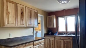 A kitchen with wooden cabinets, a black countertop, a refrigerator, and a window providing natural light. - Walker's Refuge- large family home, close to river trails (Cody)