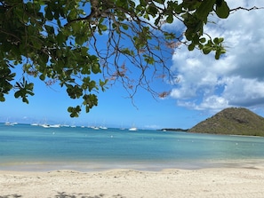 A beach with clear blue water, boats, and a hill in the background.
