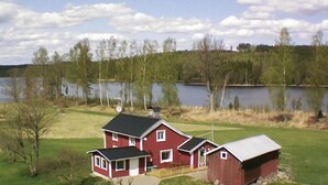 Ein rotes Haus mit schwarzem Dach, eine kleinere angebaute Garage und ein Gartenhaus, umgeben von einer grünen Wiese und einem See.