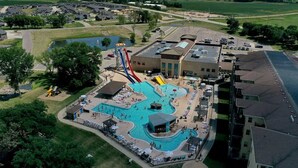 Una grande piscina con uno scivolo acquatico, circondata da lettini e ombrelloni, con un parcheggio e edifici sullo sfondo.