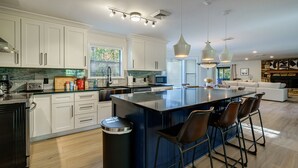 A modern kitchen with white cabinets, a large island with bar stools, a built-in oven, and a stone backsplash. - Lake Front Mountain View w/ 6 King beds (Gainesville)