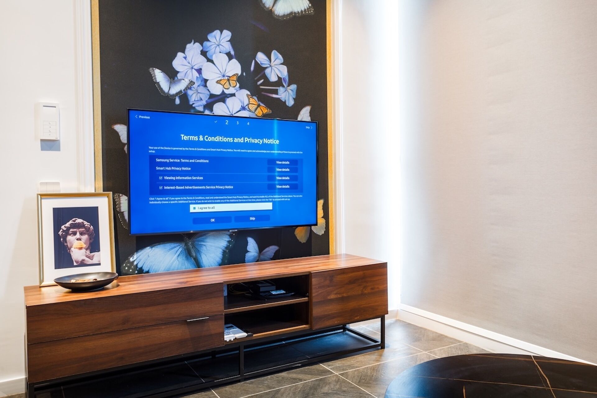A modern living room with a flat-screen TV on a wooden stand, a framed picture on the stand, and a decorative wall with flowers and butterflies.